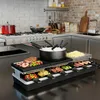 A raclette grill set is displayed on a kitchen counter, with food items and cooking utensils arranged around it. The background shows a kitchen setting with appliances and cabinetry. Náhľad obrázku produktu