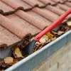 A red and black gutter cleaning tool is shown clearing leaves from a gutter on a tiled roof. Produktbild-Vorschau