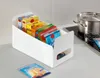A white organizer bin filled with colorful food product boxes and pouches sits on a kitchen counter next to a pot and whisk. Produktbild-Vorschau