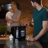A man is operating a baby bottle maker while a woman holds a baby in the background. Podgląd zdjęcia produktu