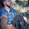 A man is sitting outdoors in a grassy area, leaning against a tree, with a backpack next to him. A red and black radio is attached to the backpack. Produktbild-Vorschau