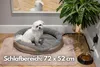 A small white dog sits in a round, heated pet bed placed on a fluffy pink rug. A plant and a dog bowl are also visible in the scene. Produktbild-Vorschau