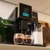 A close-up shot of a black coffee machine brewing coffee into two glass cups, with a milk container next to it. Produktbild-Vorschau