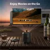 A portable projector is set up outdoors on a wooden table, projecting a movie onto a screen attached to a camper. Two bottles of alcohol and a glass of whiskey are also on the table. Náhled obrázku produktu