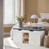 A bedroom scene with a white storage bench in the foreground, a bed and window in the background. Produktbild-Vorschau