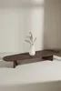 A dark brown, oval-shaped coffee table with a white vase containing dried flowers placed in the center, set against a light-colored wall with soft shadows. Produktbild-Vorschau
