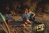 A couple having a picnic by a lake with a bicycle and a fire pit in the foreground. Produktbild-Vorschau