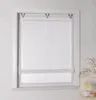 A white roller blind with a subtle pattern is installed in a window frame. A small lamp and some decorative branches are visible on the right side. Produktbild-Vorschau