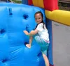 A young girl is climbing on a blue inflatable wall, which is part of a larger inflatable structure with yellow and red accents. The background shows a bright, sunny day with some greenery and buildings. Náhled obrázku produktu