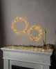 Two illuminated wreath-like light decorations are hanging on a wall above a fireplace mantelpiece, which is adorned with a garland of greenery and lights, and a vase with a plant. Podgląd zdjęcia produktu