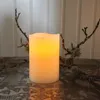 A close-up shot of a white LED candle with a warm yellow glow, placed on a decorative tray. Dried branches with lichen are arranged around the candle, set against a white paneled background. Podgląd zdjęcia produktu