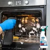 A person wearing a blue glove is spraying oven cleaner inside a dirty oven. A bottle of oven cleaner is also visible. Podgląd zdjęcia produktu