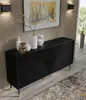 A black sideboard with decorative front panels and gold legs, placed in a room with a framed picture on the wall and a potted plant visible through a window. Produktbild-Vorschau