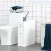 A white bathroom storage unit with two drawers is placed next to a toilet. The unit has silver handles and a small glass cup with purple flowers on top. A black wire basket with folded blue towels sits on top of the toilet tank. A white woven basket with a Voir les images du produit