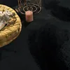 A close-up view of a black fluffy rug with a yellow cushion, a book, a candle, and a gold wire basket. Produktbild-Vorschau
