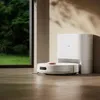 A white robot vacuum cleaner is docked next to its charging station in a room with a large window overlooking a green garden. Anteprima dell’immagine prodotto