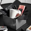 A white mug and a chocolate bar are presented in a gift box lined with black tissue paper. Produktbild-Vorschau