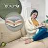 A woman is sitting on a couch reading a book while wrapped in a beige blanket. An infographic shows the layers of the blanket. Produktbild-Vorschau