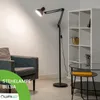 A modern black floor lamp is positioned next to a gray armchair and a white shelving unit in a living room setting. Produktbild-Vorschau