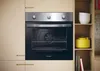 A built-in oven is shown in a kitchen setting, with shelves to the right containing kitchenware and books. Voir les images du produit