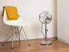 A white chair with yellow pants draped over it, next to a silver stand fan. Produktbild-Vorschau