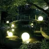 A serene garden scene at dusk, illuminated by glowing spherical lights placed around a pond and a wooden bridge. Náhľad obrázku produktu