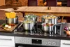 A kitchen scene with pots and pans on a stovetop, a dining table in the background. Produktbild-Vorschau