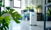 A portable air conditioner is shown in a modern office or living space with plants and furniture. Podgląd zdjęcia produktu