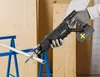 A person wearing gloves is using a black reciprocating saw to cut a metal pipe supported by a blue stand. The background consists of wooden boards. Produktbild-Vorschau