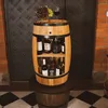 A wooden barrel-shaped wine cabinet with shelves filled with bottles and a bowl of grapes and a glass of wine on top, placed on a tiled floor. Produktbild-Vorschau