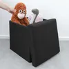 A hand is placing a stuffed monkey into a black storage ottoman. A stuffed dinosaur is also visible inside the ottoman. Náhled obrázku produktu