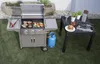 A gas grill with food cooking on it, a propane tank, and a small table with food and utensils set up in a backyard setting. Náhled obrázku produktu