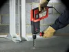 A person wearing gloves is using a Black&Decker rotary hammer drill to drill into a concrete floor next to some white support beams. Produktbild-Vorschau