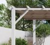 A close-up view of a white pergola with a light brown canopy, set against a backdrop of green trees and a white wall. Podgląd zdjęcia produktu