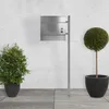 A modern stainless steel mailbox stand with a mailbox attached, placed on a tiled patio next to two potted plants. Náhled obrázku produktu