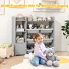 A young girl is sitting on the floor in front of a toy shelf, holding a teddy bear. The shelf is filled with toys and storage bins. Podgląd zdjęcia produktu