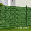 A green fence with a leaf pattern, with a building in the background and grass in the foreground. Náhled obrázku produktu