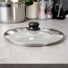A glass pot lid with a black handle rests on a speckled gray countertop, with a stainless steel pot and kitchen accessories blurred in the background. Náhľad obrázku produktu