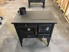 A black wood-burning stove with a metal cooktop and oven is displayed in a warehouse or workshop setting. Anteprima dell’immagine prodotto