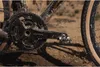 Close-up of a muddy bicycle crankset and pedal, with a tire visible on the right, on a dirt surface. Produktbild-Vorschau