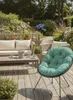 A comfortable outdoor seating area with a sofa, coffee table, and a round chair with a green cushion, surrounded by lush garden plants and flowers. Produktbild-Vorschau
