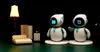 Two small robots are positioned in a miniature bedroom setting with a bed, wardrobe, and nightstand. A lamp is also present. Náhled obrázku produktu
