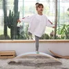 A young girl is balancing on a wooden balance board in a bright room with large windows and plants. Produktbild-Vorschau