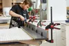 A person is using a drill to attach a piece of wood to another piece of wood, which is held in place by several clamps. The clamps are black and red. Náhľad obrázku produktu
