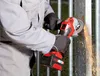 A person wearing work clothes and gloves is using a red and black cordless angle grinder to cut a metal fence post, creating a shower of sparks. Podgląd zdjęcia produktu
