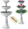 Two white tiered plant stands are shown against a white background. One is empty and the other is filled with colorful flowers. Produktbild-Vorschau