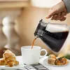 A person is pouring coffee from a carafe into a white mug, with croissants and pastries on a table. Produktbild-Vorschau