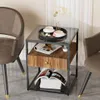 A small side table with a glass top, a drawer, and a shelf, placed between two chairs in a room with sheer curtains in the background. Produktbild-Vorschau