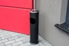 A black cylindrical trash can with an ashtray top stands on a paved surface next to a red wall and a gray building. Náhled obrázku produktu