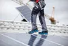 A person wearing safety shoes and work pants walks on a rooftop with solar panels, holding a rope. A construction crane is visible in the background. Náhľad obrázku produktu